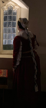 A picture of Isabel Maria, from behind, as she looks out a gothic revivial window while wearing a cofia y tranzada, a red velvet gonete (trimmed in gold lace) with a gold-work belt, worn over a red linen skirt with three horizontal pleats, between courses during the An Evening in Granada event